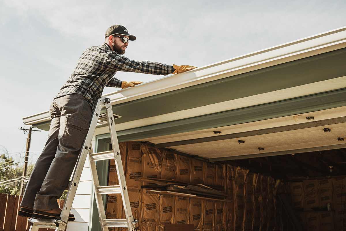 A man cleans out his gutters while standing on a ladder
